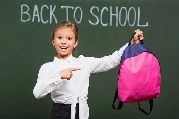 Foco seletivo de colegial sorridente apontando para mochila rosa perto de volta para a escola lettering no quadro — Fotografia de Stock