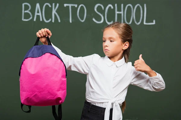 Linda colegiala mostrando el pulgar hacia arriba mientras sostiene la mochila cerca de pizarra con letras de vuelta a la escuela - foto de stock