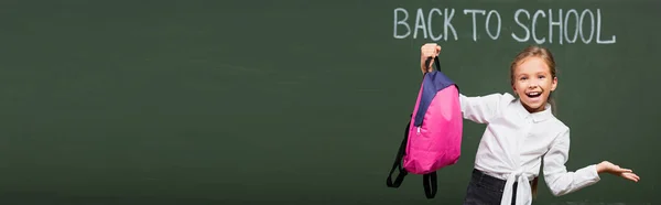 Imagen horizontal de colegiala alegre con brazo abierto sosteniendo la mochila cerca de pizarra con inscripción de regreso a la escuela - foto de stock