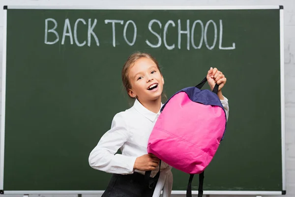 Scolaretta felice mostrando zaino rosa vicino alla lavagna con iscrizione a scuola — Foto stock