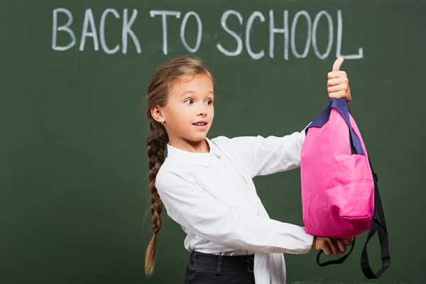 Surprised schoolgirl holding pink backpack near chalkboard with back to school lettering — Stock Photo