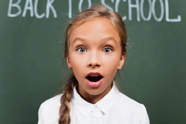 Selective focus of shocked schoolgirl with open mouth looking at camera near chalkboard with back to school lettering — Stock Photo