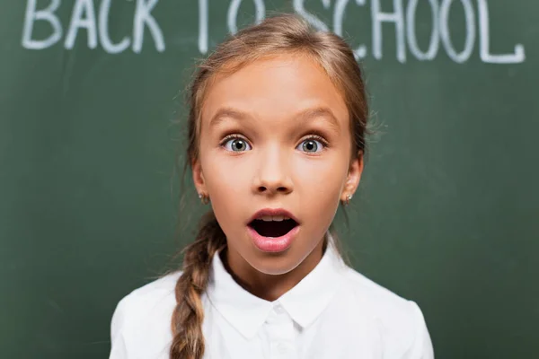 Messa a fuoco selettiva della studentessa scioccata con la bocca aperta guardando la fotocamera vicino alla lavagna con la scritta di ritorno a scuola — Foto stock