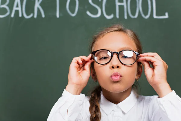 Selective focus of cute schoolgirl touching eyeglasses and blowing air kiss near chalkboard with back to school lettering — Stock Photo