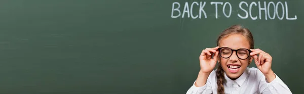 Horizontal image of excited schoolgirl laughing with closed eyes and touching eyeglasses near chalkboard with back to school text — Stock Photo