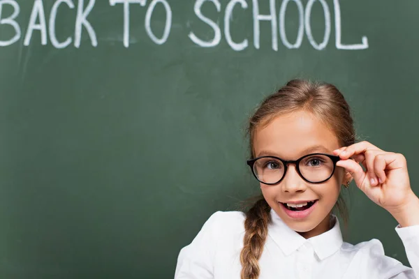 Messa a fuoco selettiva di scolara allegra che tocca occhiali vicino a lavagna con ritorno a iscrizione scolastica — Foto stock