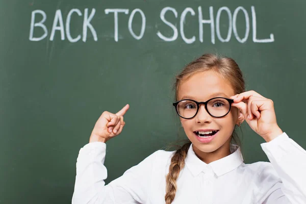 Selective focus of smiling schoolgirl touching eyeglasses and pointing with finger at chalkboard with back to school lettering — Stock Photo