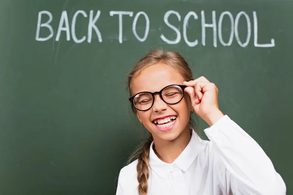Studentessa eccitata ridendo con gli occhi chiusi e toccando gli occhiali vicino alla lavagna con la scritta torna a scuola — Foto stock