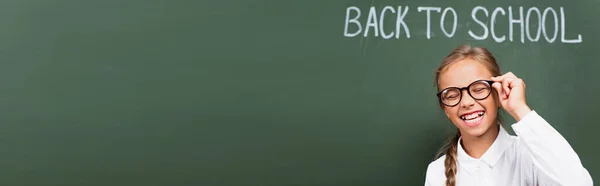 Panoramic crop of excited schoolgirl laughing with closed eyes and touching eyeglasses near chalkboard with back to school lettering — Stock Photo