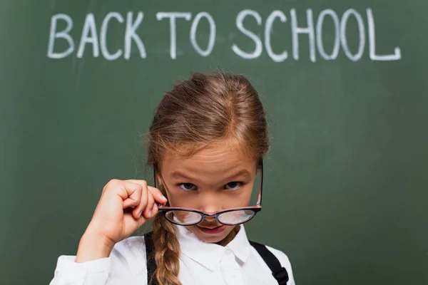 Foco seletivo de colegial bonito decolando óculos enquanto olha para a câmera perto de volta para a escola lettering no quadro — Fotografia de Stock