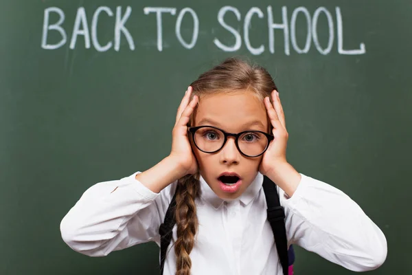 Selective focus of shocked schoolgirl in eyeglasses touching head near back to school lettering on chalkboard — Stock Photo