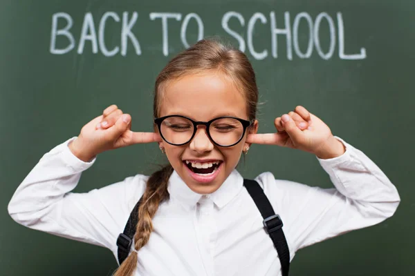 Selective focus of displeased schoolgirl in eyeglasses plugging ears with fingers near back to school inscription on chalkboard — Stock Photo
