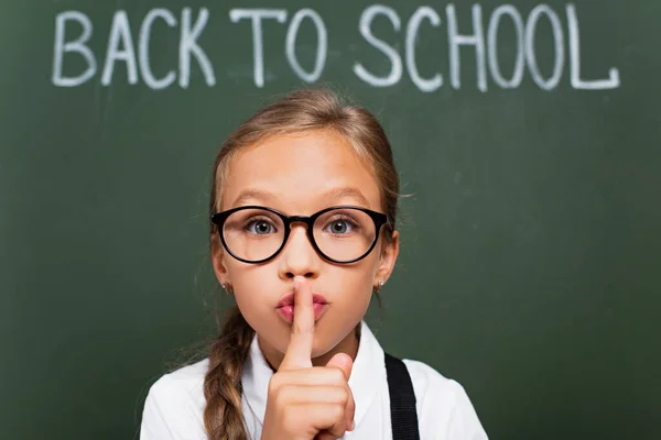 Selektiver Fokus des netten Schulmädchens in Brille, das Schweigezeichen in der Nähe der Tafel mit der Schulaufschrift zeigt — Stockfoto