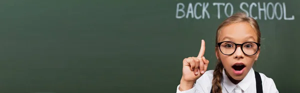 Image horizontale d'une écolière surprise montrant un geste d'idée près d'un tableau noir avec un lettrage de retour à l'école — Photo de stock