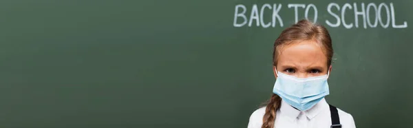 Imagen horizontal de colegiala disgustada en máscara protectora mirando a la cámara cerca de la inscripción de la escuela en pizarra - foto de stock