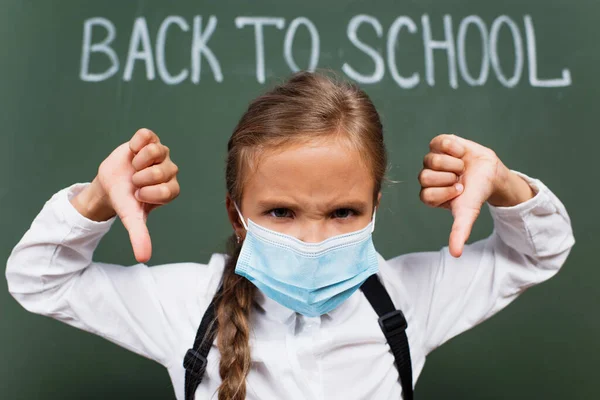 Selective focus of displeased schoolgirl in medical mask showing thumbs down near chalkboard with back to school inscription — Stock Photo