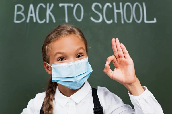 Enfoque selectivo de colegiala positiva en máscara médica mostrando gesto bien cerca de la inscripción de la escuela en pizarra - foto de stock