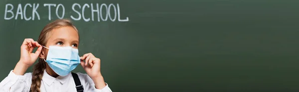 Conceito panorâmico de estudante colocando máscara médica perto de volta à inscrição da escola no quadro — Fotografia de Stock