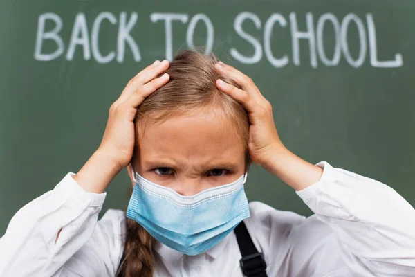 Foco seletivo de franzir a testa, menina exausta em máscara médica tocando cabeça perto de volta para a escola lettering no quadro — Fotografia de Stock