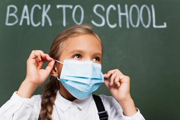 Enfoque selectivo de la colegiala que se pone la máscara médica cerca de la letra de la escuela en pizarra - foto de stock
