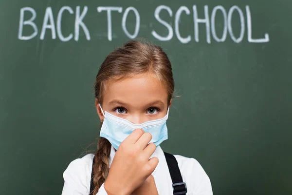 Foyer sélectif d'écolière mignonne touchant masque médical et regardant la caméra près de tableau noir avec inscription de retour à l'école — Photo de stock