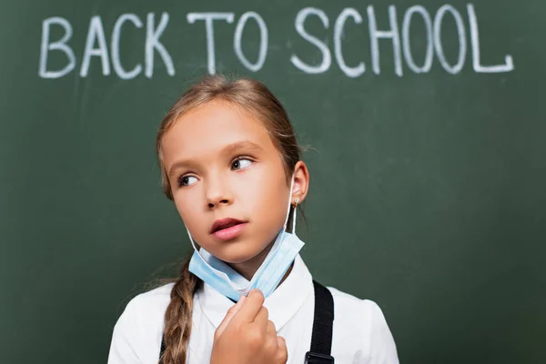 Selective focus of exhausted schoolgirl taking off protective mask near chalkboard with back to school lettering — Stock Photo