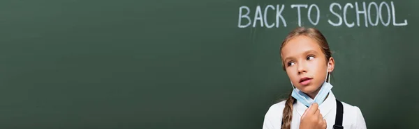 Horizontal concept of tired schoolgirl taking off protective mask near chalkboard with back to school lettering — Stock Photo