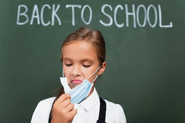 Foyer sélectif d'écolière mécontente touchant masque de protection près de tableau noir avec texte de retour à l'école — Photo de stock