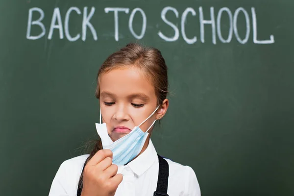 Messa a fuoco selettiva di studentessa scontento toccando maschera protettiva vicino lavagna con torna al testo della scuola — Foto stock