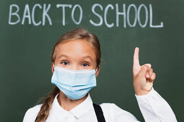 Selective focus of cute schoolgirl in protective mask looking at camera near chalkboard with back to school lettering — Stock Photo