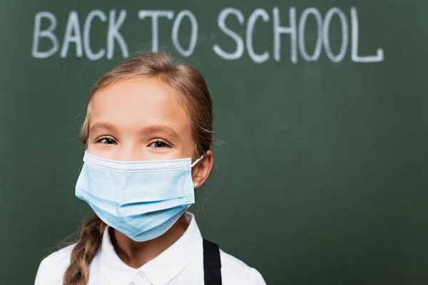 Enfoque selectivo de colegiala positiva en máscara médica mirando a la cámara cerca de pizarra con inscripción de vuelta a la escuela - foto de stock