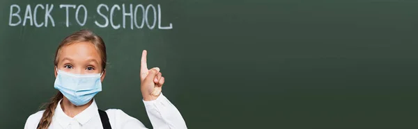 Imagen horizontal de colegiala en máscara médica apuntando con el dedo cerca de pizarra con inscripción de vuelta a la escuela - foto de stock