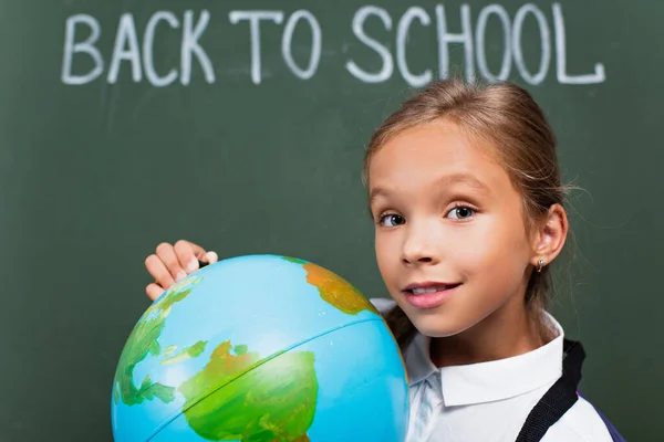 Selective focus of smiling schoolgirl holding globe and looking at camera near back to school inscription on chalkboard — Stock Photo