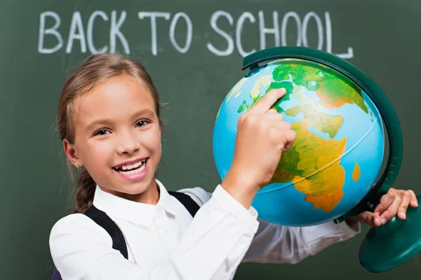 Enfoque selectivo de colegiala feliz señalando con el dedo en el globo cerca de la inscripción de la escuela en pizarra - foto de stock