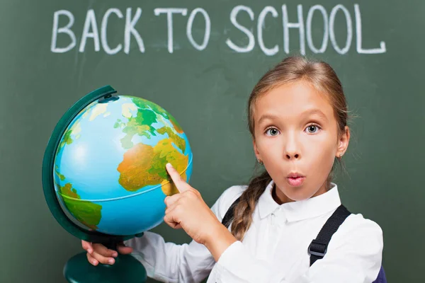 Colegiala sorprendida señalando con el dedo en el globo cerca de la letra de la escuela en pizarra - foto de stock