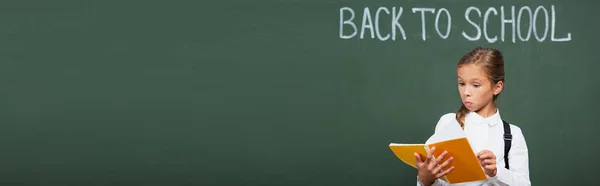 Horizontal image of surprised schoolgirl looking at copy book near chalkboard with back to school lettering — Stock Photo