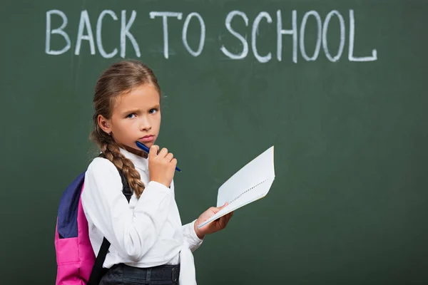 Upset schoolgirl with backpack holding notebook and pen near chalkboard with back to school lettering — Stock Photo