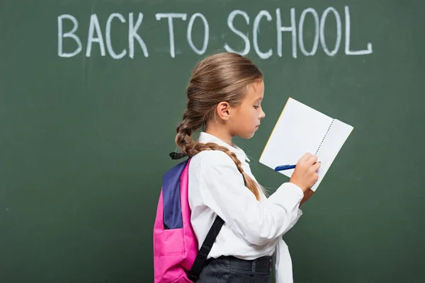 Écolière avec sac à dos écrivant dans le livre de copie près de tableau noir avec inscription de retour à l'école — Photo de stock