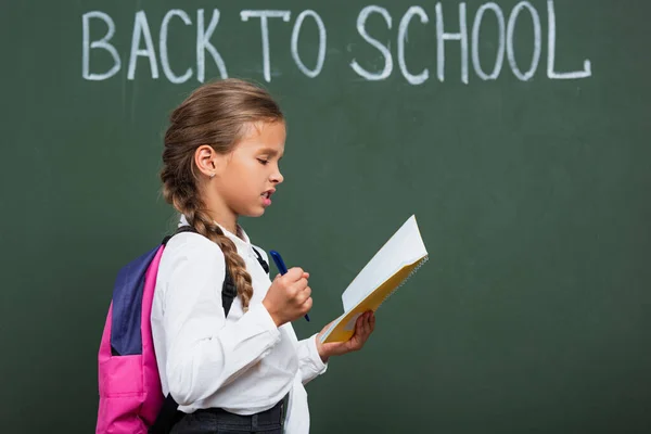 Vista laterale della scolaretta con zaino che tiene quaderno e penna vicino alla lavagna con iscrizione a scuola — Foto stock