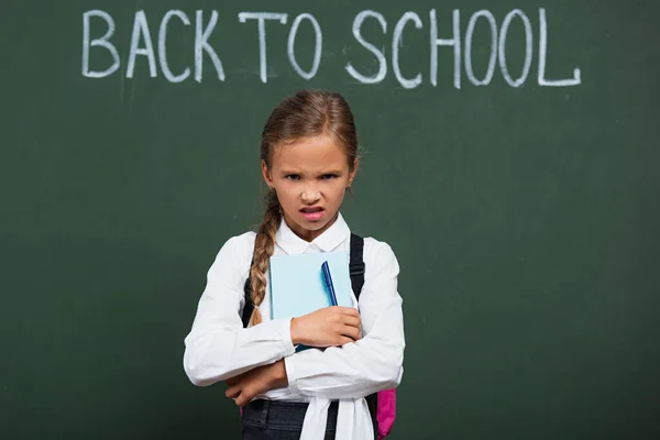 Mécontent écolière tenant stylo et livre près de tableau noir avec retour à l'école lettrage — Photo de stock