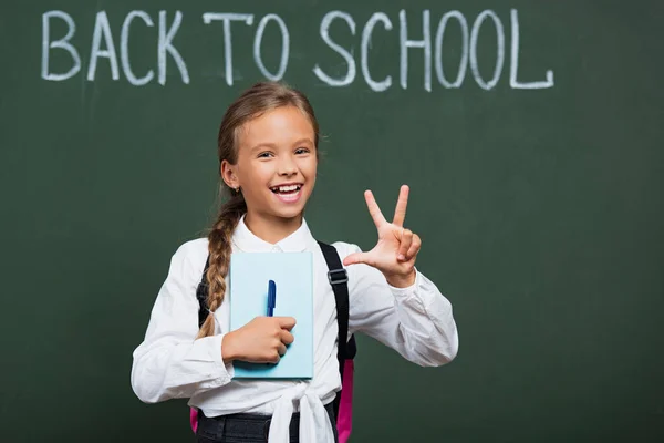 Scolaretta felice con libro e penna che mostra il gesto della vittoria vicino alla lavagna con testo di ritorno a scuola — Foto stock