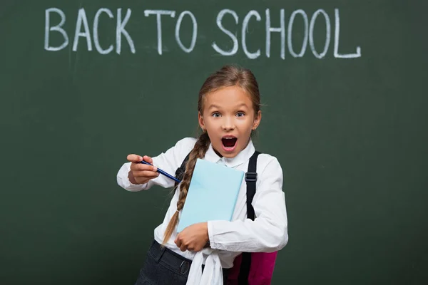 Estudante animado apontando com caneta no livro perto de quadro-negro com volta para a escola lettering — Fotografia de Stock