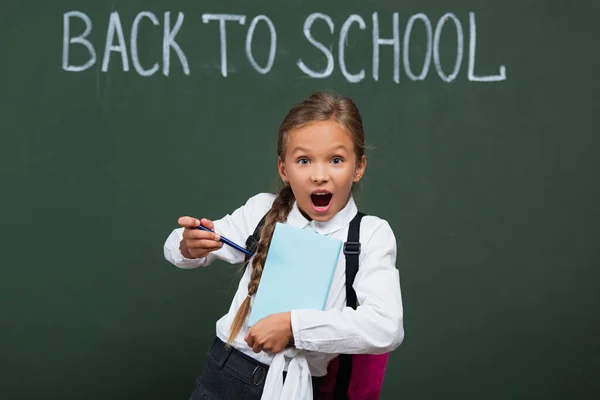 Colegiala emocionada señalando con pluma en el libro cerca de pizarra con letras de vuelta a la escuela - foto de stock