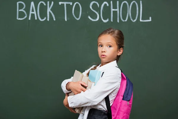 Écolière découragée avec sac à dos tenant des livres près du tableau noir avec inscription de retour à l'école — Photo de stock