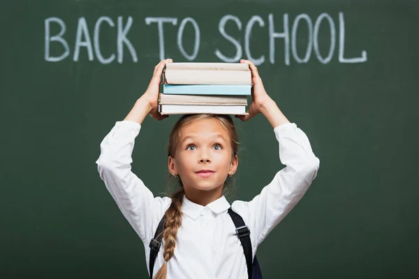 Positive schoolgirl holding stack of books above head near chalkboard with back to school lettering — Stock Photo