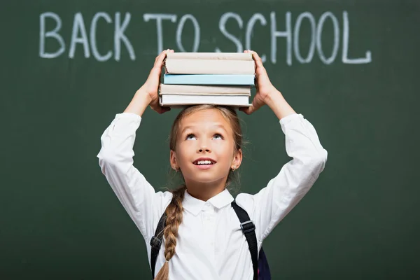 Enfoque selectivo de la colegiala sonriente sosteniendo pila de libros por encima de la cabeza cerca de pizarra con texto de vuelta a la escuela - foto de stock