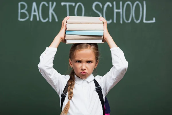 Selective focus of displeased schoolgirl holding stack of books above head near chalkboard with back to school lettering — Stock Photo