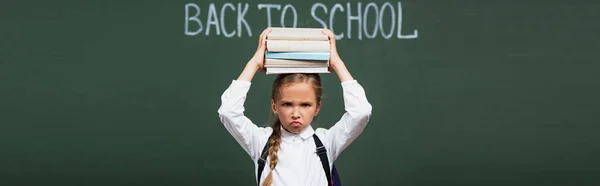 Récolte panoramique d'écolière mécontente tenant pile de livres au-dessus de la tête près du tableau noir avec lettrage de retour à l'école — Photo de stock