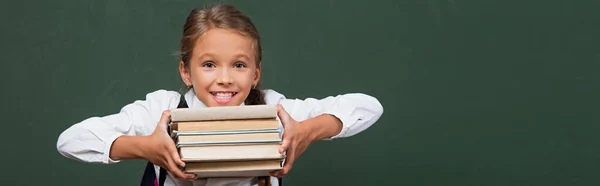 Imagen horizontal de colegiala feliz mostrando pila de libros cerca de pizarra verde - foto de stock