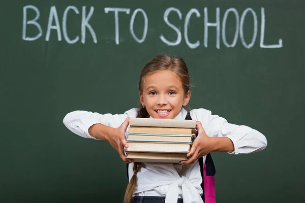 Excited schoolgirl holding stack of books near chalkboard with back to school inscription — Stock Photo